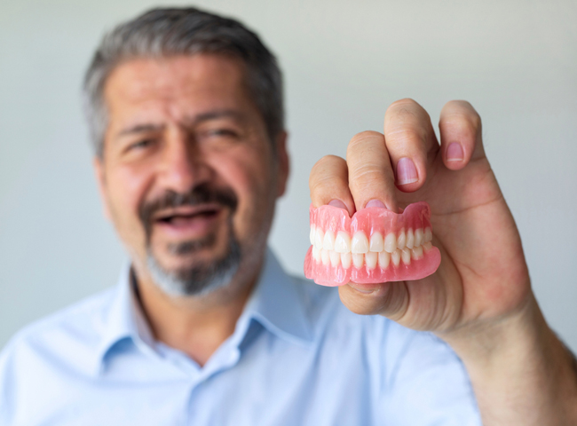Dentist holding pink dentures, smiling in clinical setting, wearing blue shirt