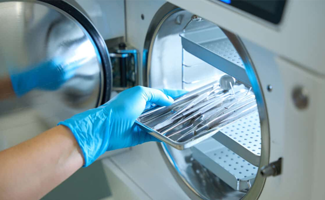 Dental professional in blue gloves sterilizing instruments in autoclave machine.