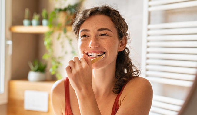 Woman smiling while brushing teeth in bright bathroom with plants and shelving.