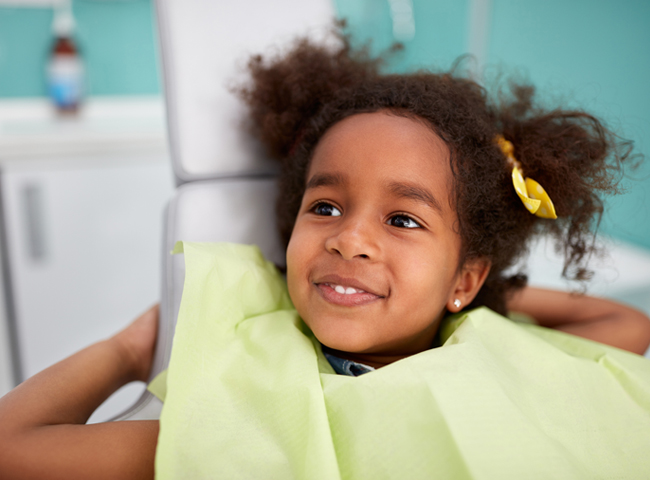 Young child smiling at dental office, wearing yellow shirt with hair clips