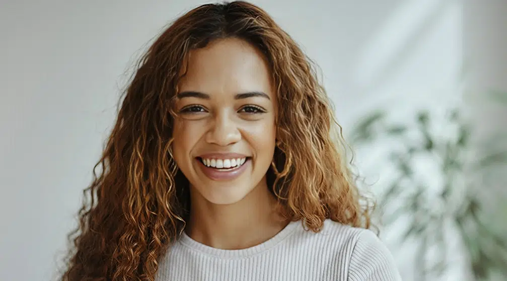Young woman with curly brown hair smiling at camera in bright dental office setting.