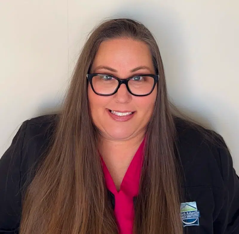 Professional headshot of smiling woman with glasses wearing pink top and black blazer against white background.